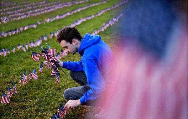 A person wearing a blue sweatshirt kneels as they place small American flags into a lawn.  